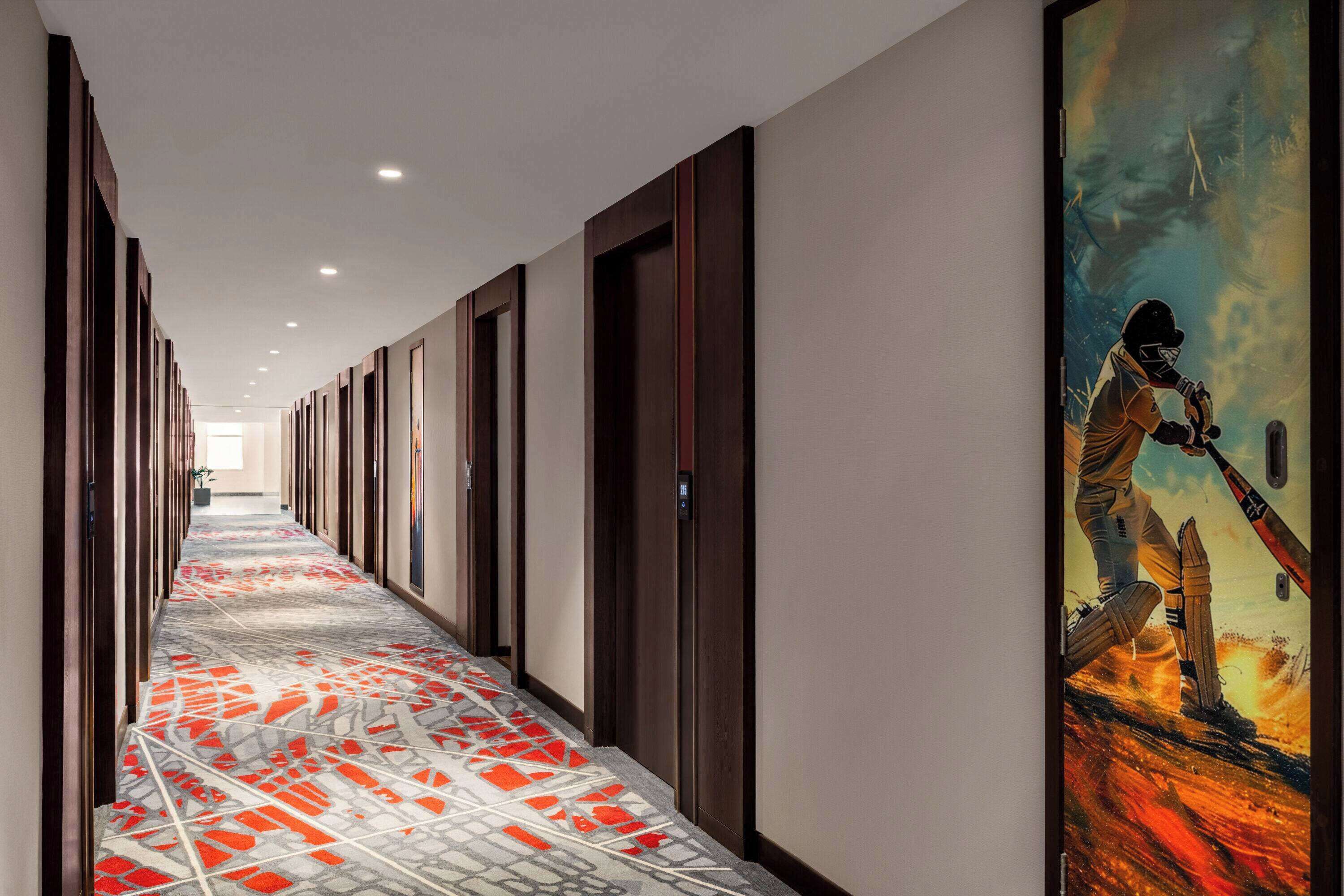 Un pasillo de hotel con piso estampado, puertas de madera y un cuadro grande en la pared derecha.