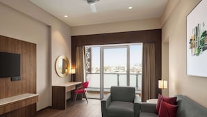 A modern hotel room with a large window offering a city view, a flat-screen TV mounted on the wall, a grey sofa with red pillows, a small round coffee table, and a desk with a red chair. - Ramada by Wyndham Ahmedabad Narendra Modi Stadium Motera (Ahmedabad)