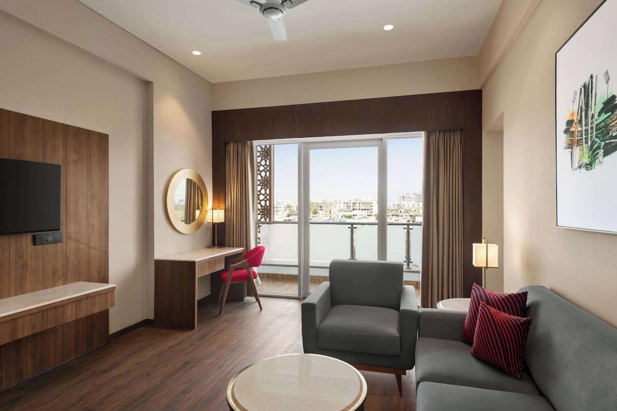 A modern hotel room with a large window offering a city view, a flat-screen TV mounted on the wall, a grey sofa with red pillows, a small round coffee table, and a desk with a red chair.