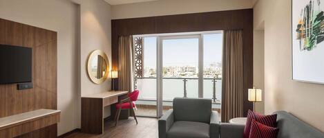 A modern hotel room with a large window offering a city view, a flat-screen TV mounted on the wall, a grey sofa with red pillows, a small round coffee table, and a desk with a red chair.