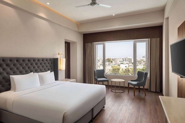 A hotel room with a large bed, two chairs, a small table, a TV, and a window with a city view. - Ramada by Wyndham Ahmedabad Narendra Modi Stadium Motera (Ahmedabad)