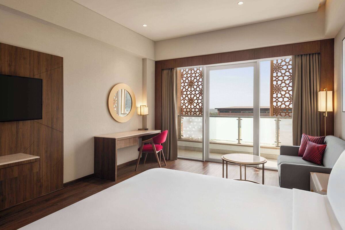 A hotel room with a large bed, a desk with a chair, a TV mounted on the wall, and a balcony with a view.