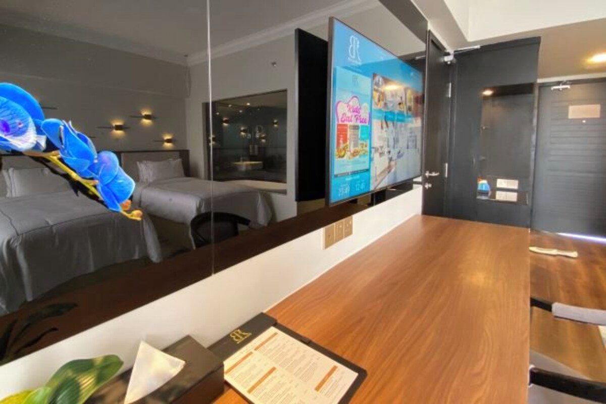 Un espace d’accueil d’hôtel avec un comptoir de réception en bois, un écran numérique affichant un menu, et un lit doté d’une tête de lit à motifs floraux bleus.