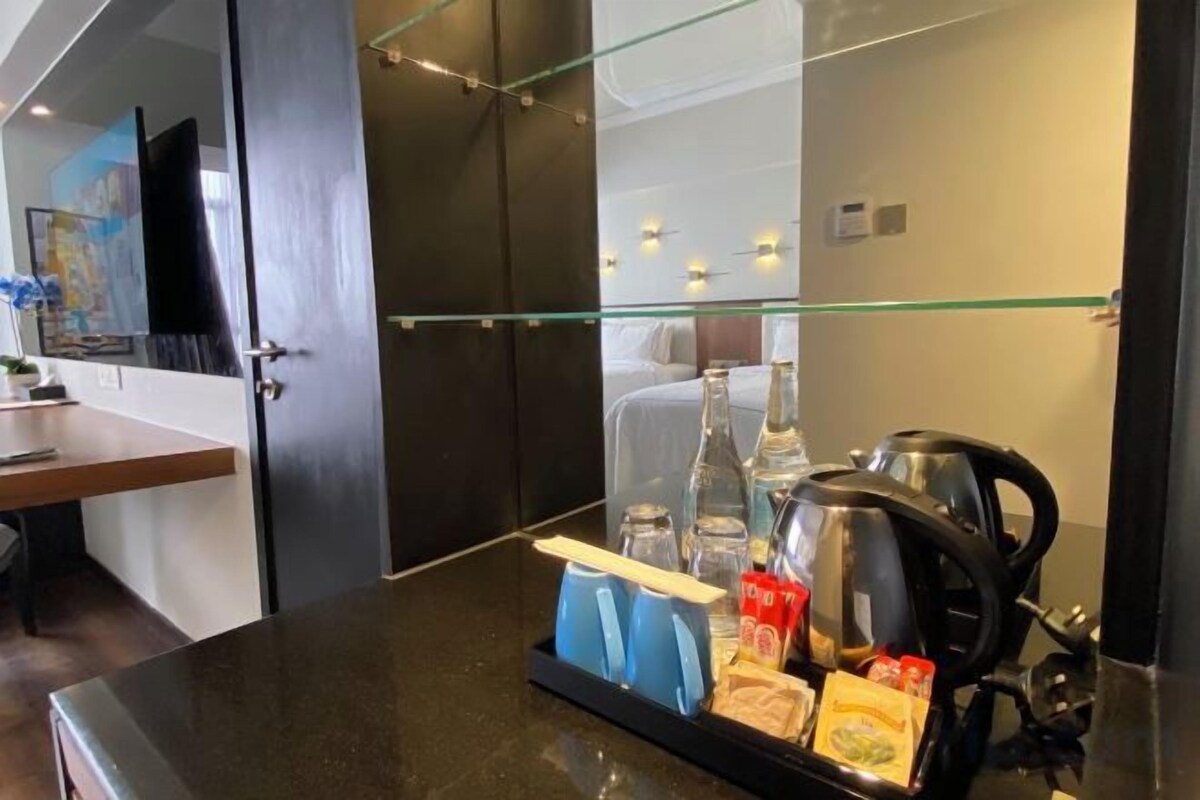 Et hotelværelse med glasafskærmning, et tekøkken med kaffemaskine og et morgenmadsserveringsfad med snacks.