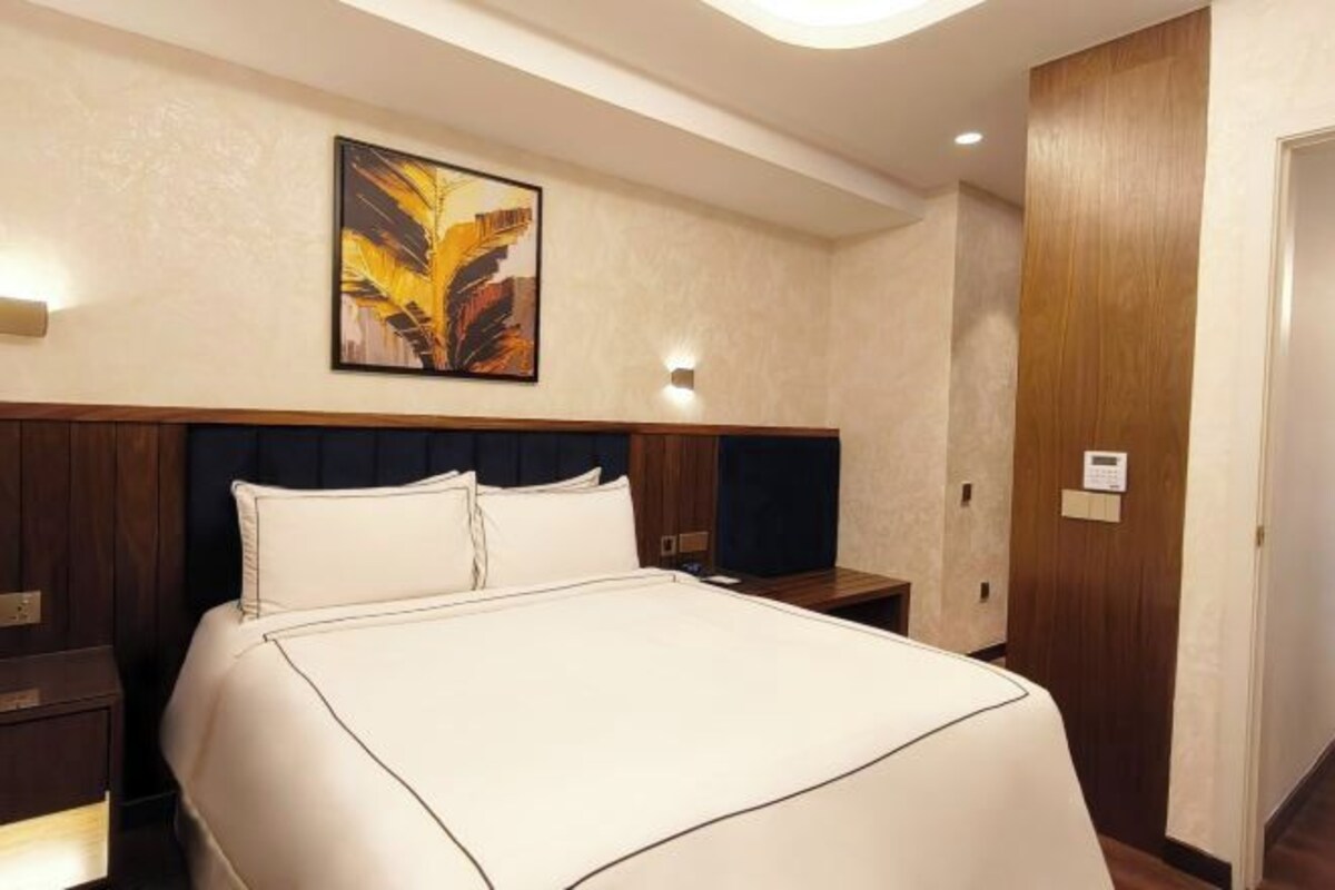 A hotel room with a large bed, a headboard with a wooden panel, a wall-mounted artwork, and a nightstand with a lamp.