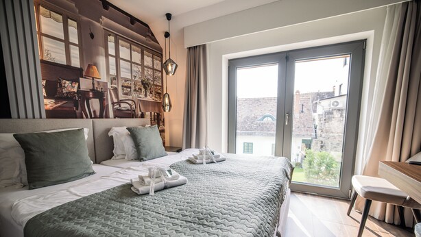 A hotel room with a large bed, a desk, and a view of the city through a large window. - Rókusfalvy Borhotel (Etyek)
