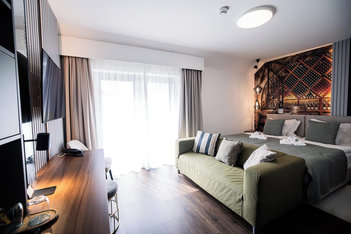Una habitación de hotel moderna con un escritorio de madera, un sofá verde, una cama con cabecera estampada y una estantería para vinos.