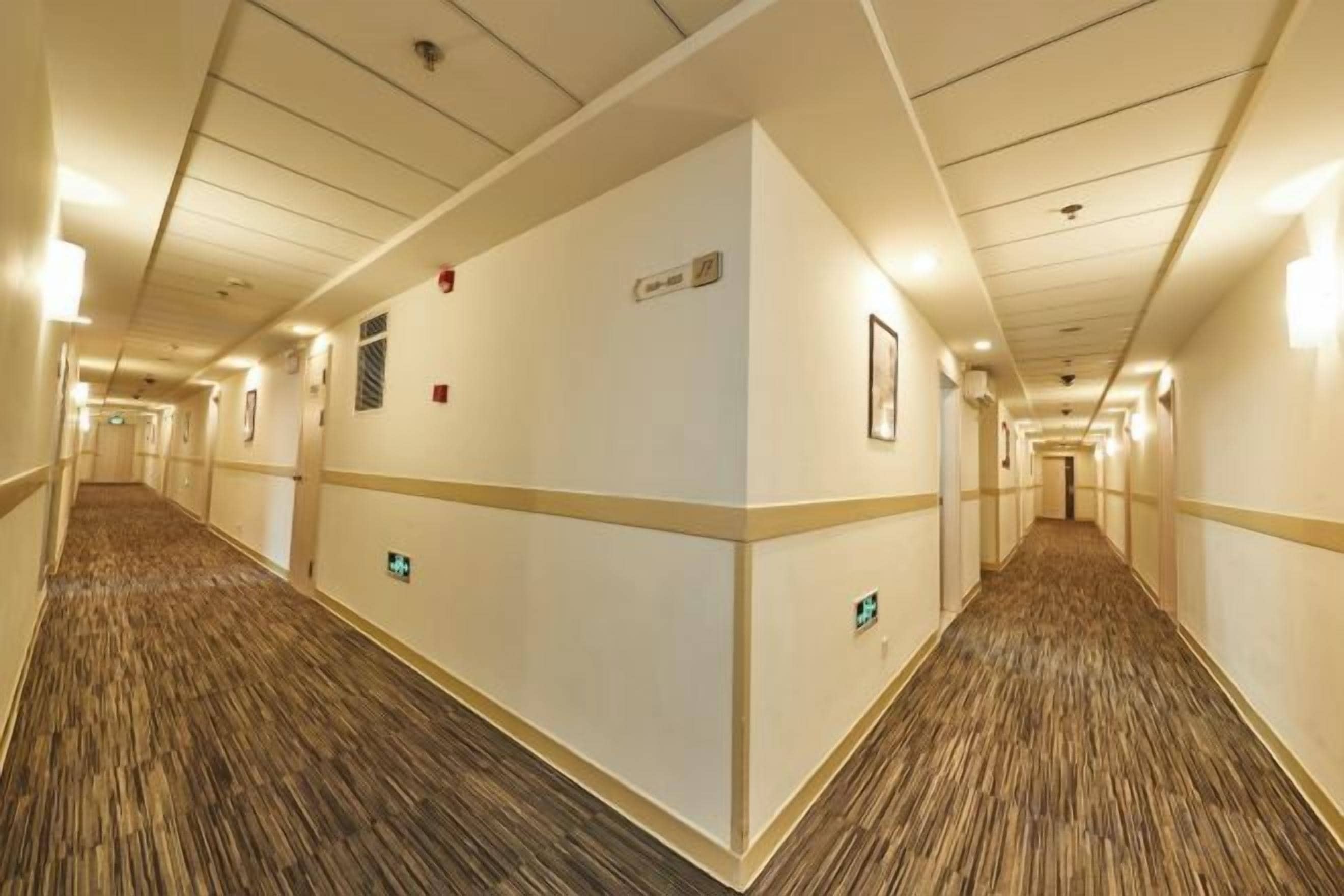 A hotel corridor with carpeted floors, walls with light fixtures, and emergency exit signs.