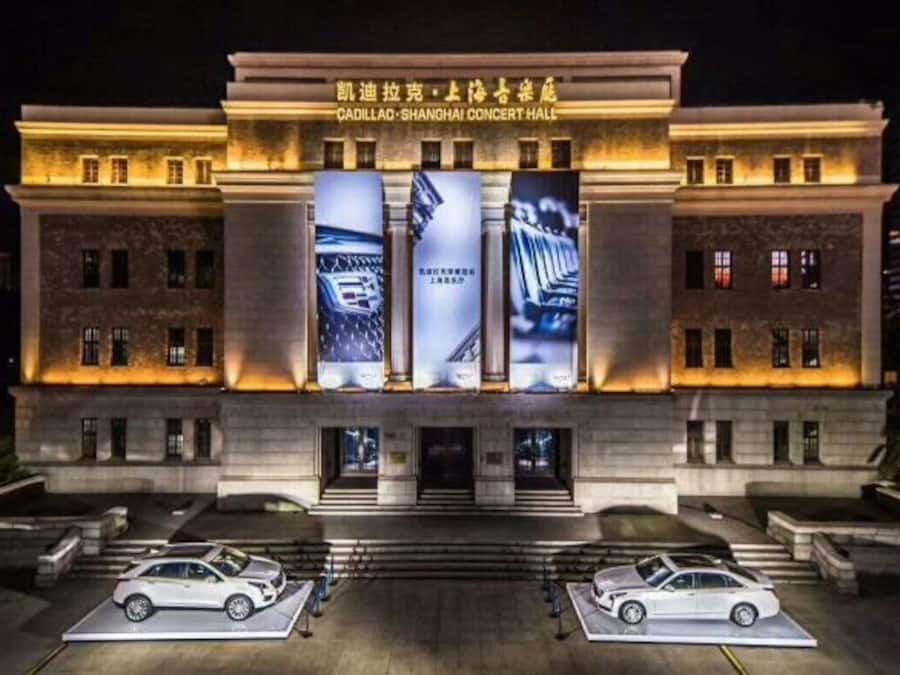 Um edifício com uma grande placa indicando 'Cadillac-Shanghai Concert Hall' e dois carros estacionados na frente.