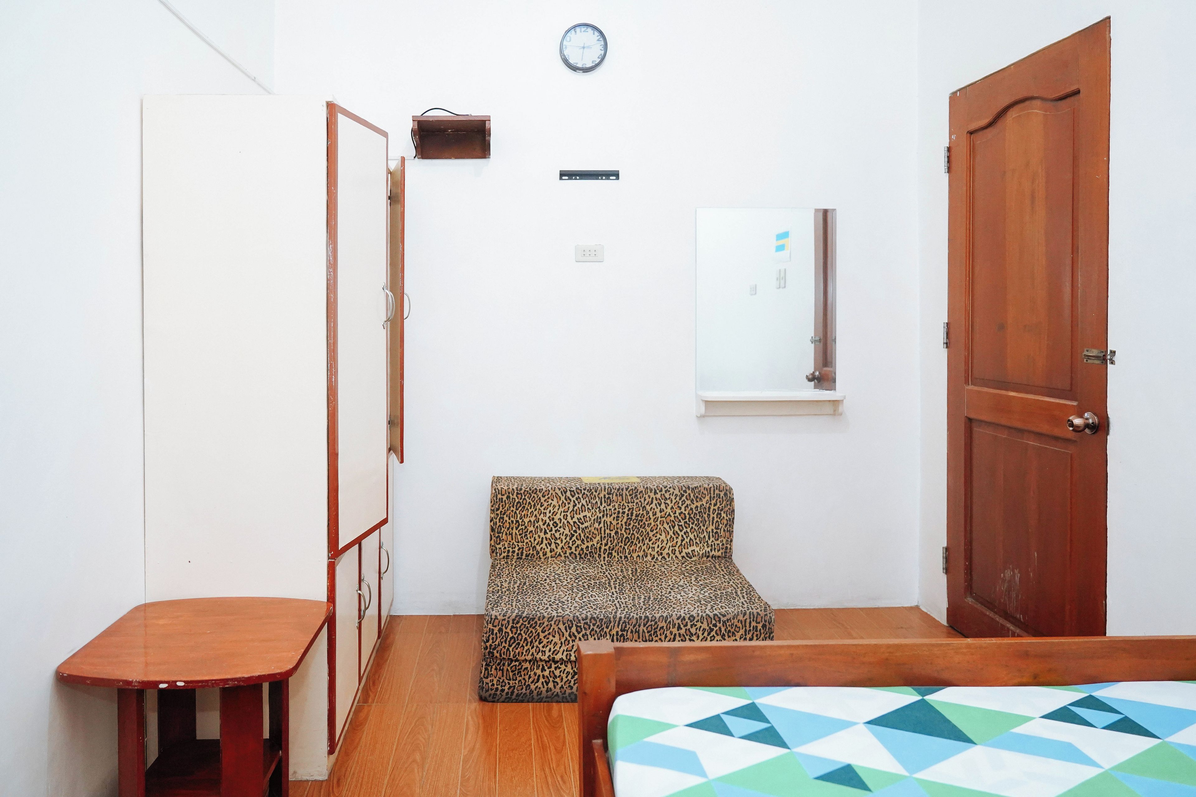 A small room with a bed, a wooden table, a leopard print chair, a wooden wardrobe, a wooden door, a wall clock, and a window.