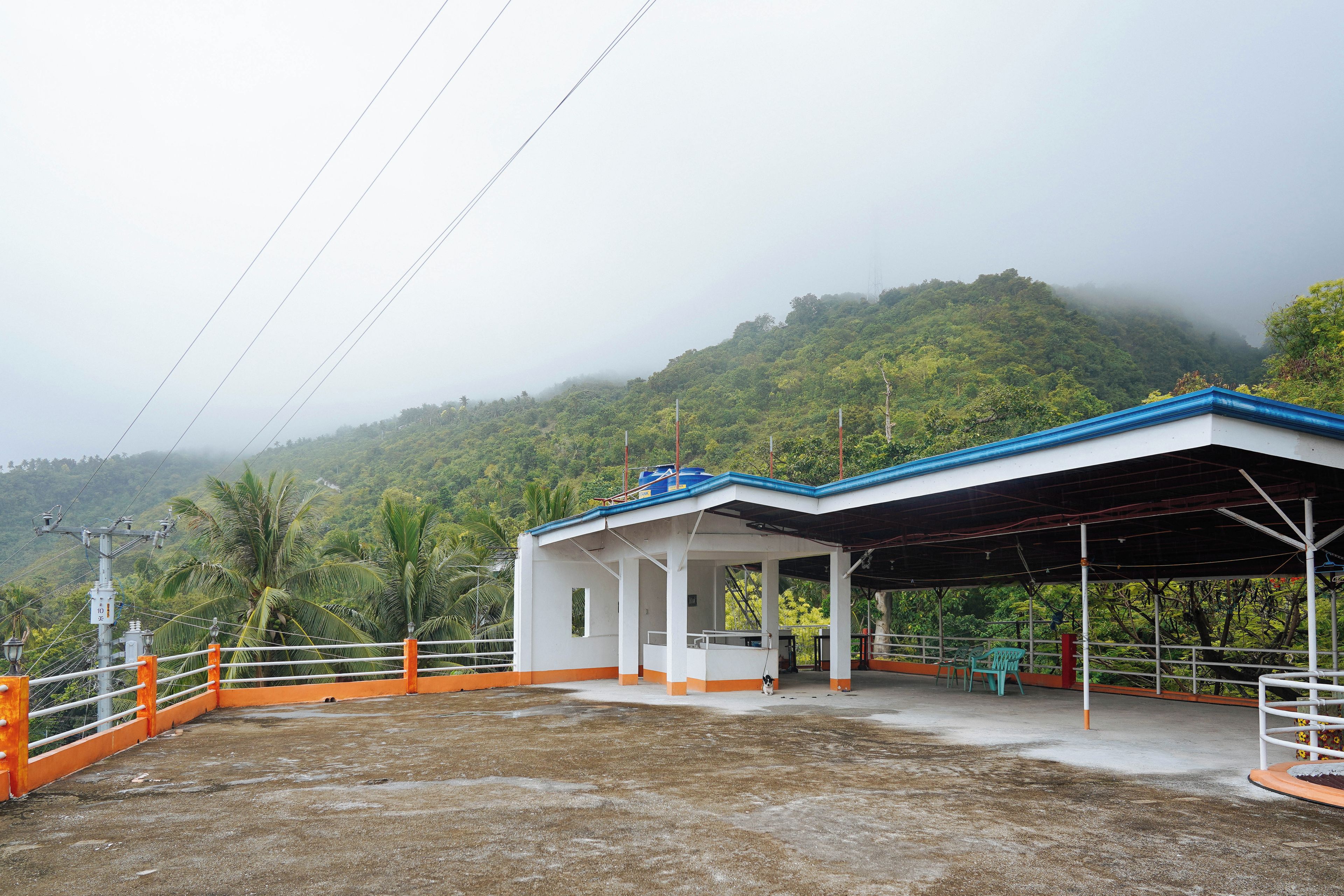 A covered outdoor area with a white building, orange railings, and a view of a forested hill.