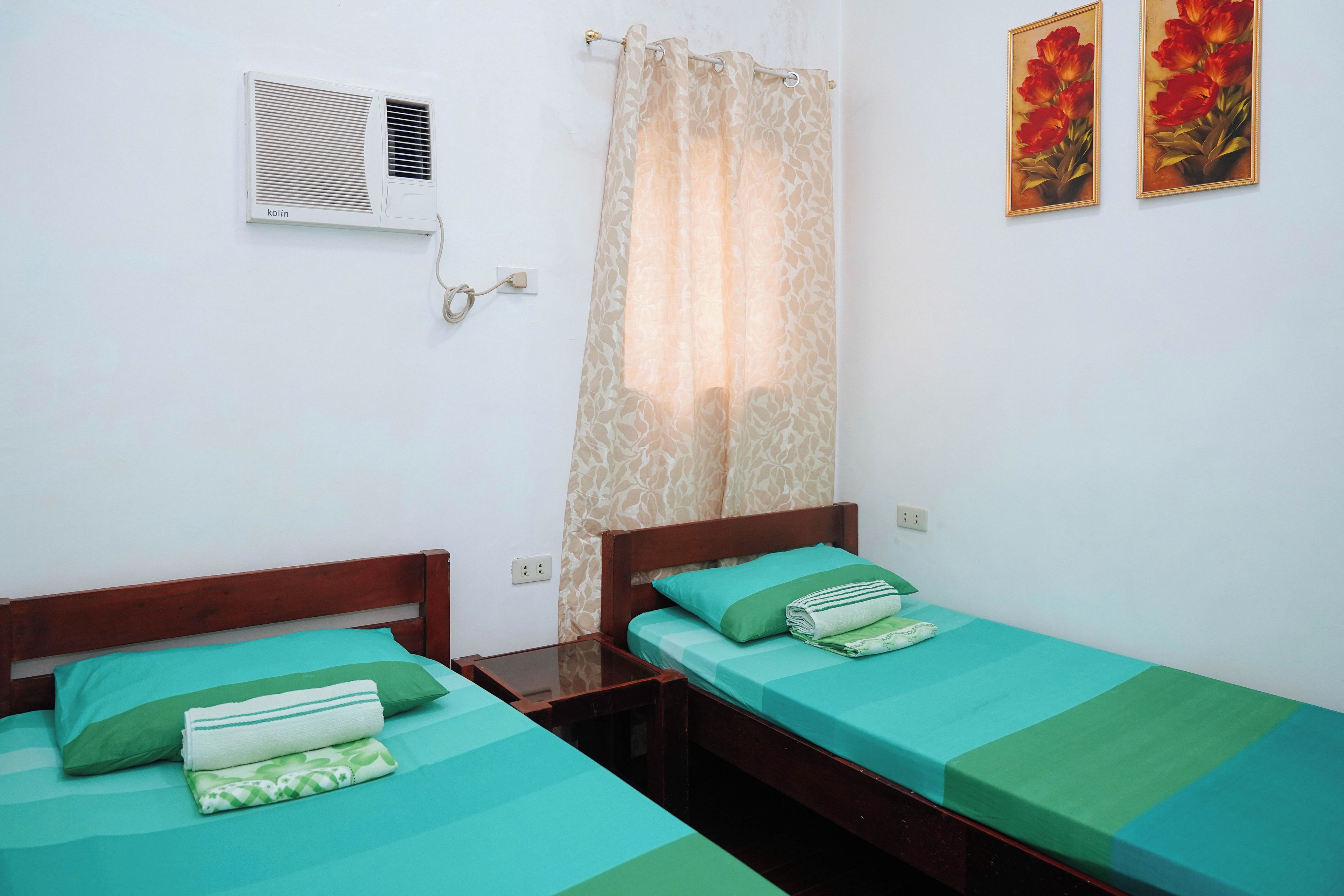 A room with two single beds, each with green bedding and pillows, a wooden headboard, a small bedside table, and a wall-mounted air conditioner.