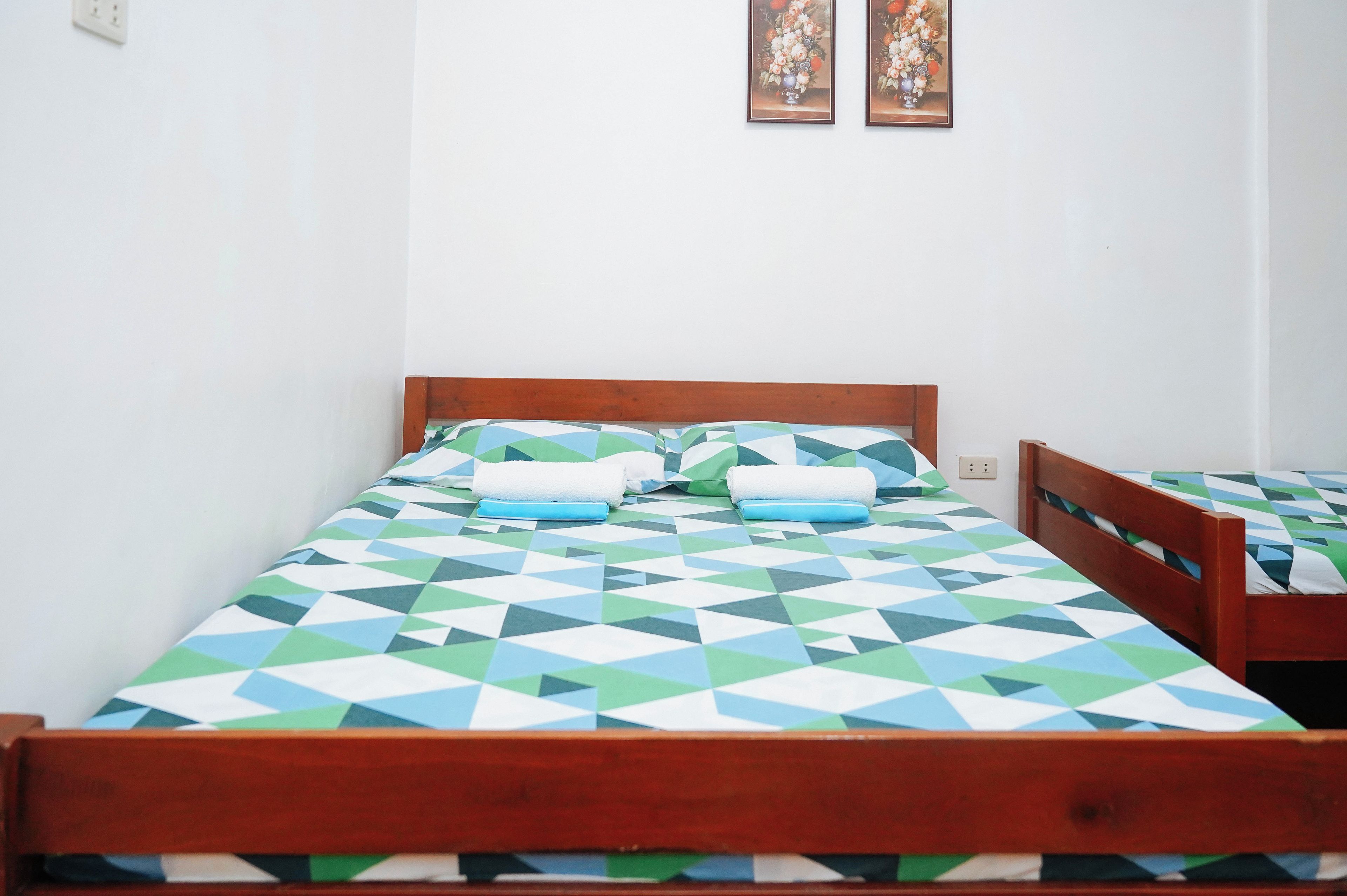 A single bed with a geometric patterned bedspread, a wooden headboard, and two framed floral paintings on the wall.