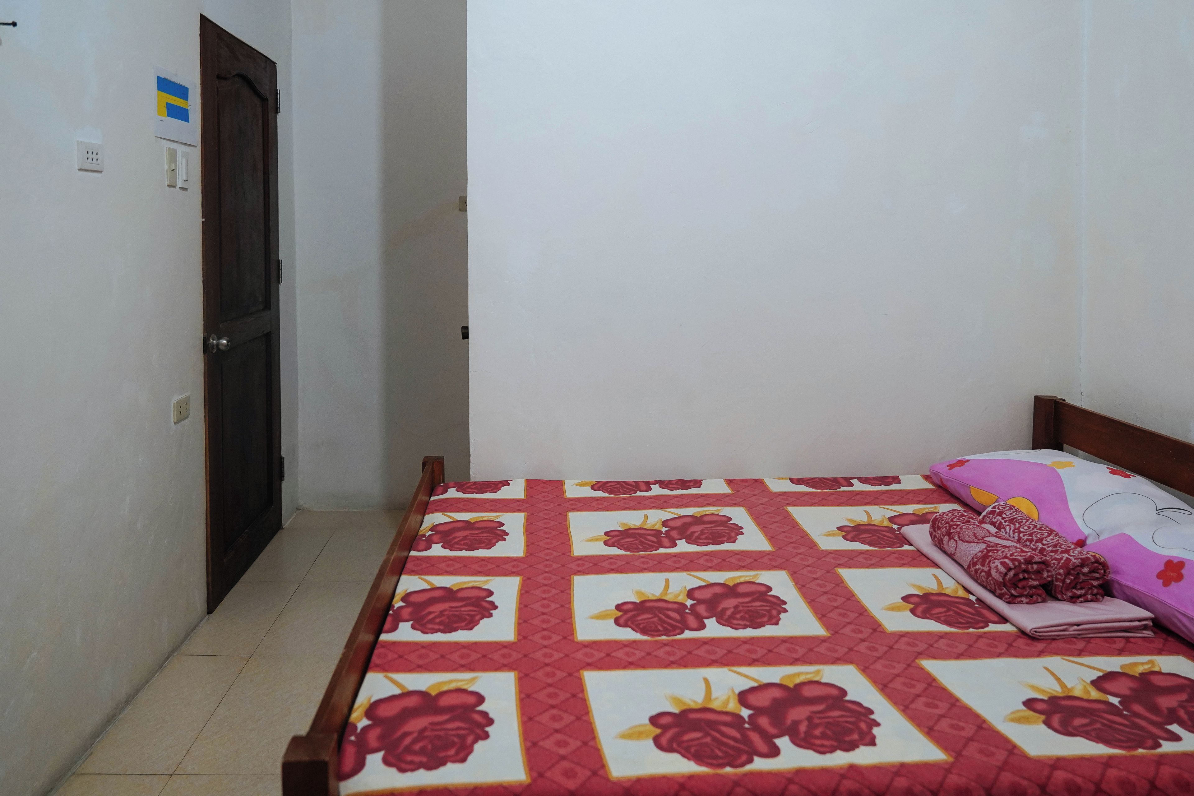 A bedroom with a bed covered in a floral-patterned bedspread, a wooden headboard, and a nightstand with a rolled-up towel and a pillow.