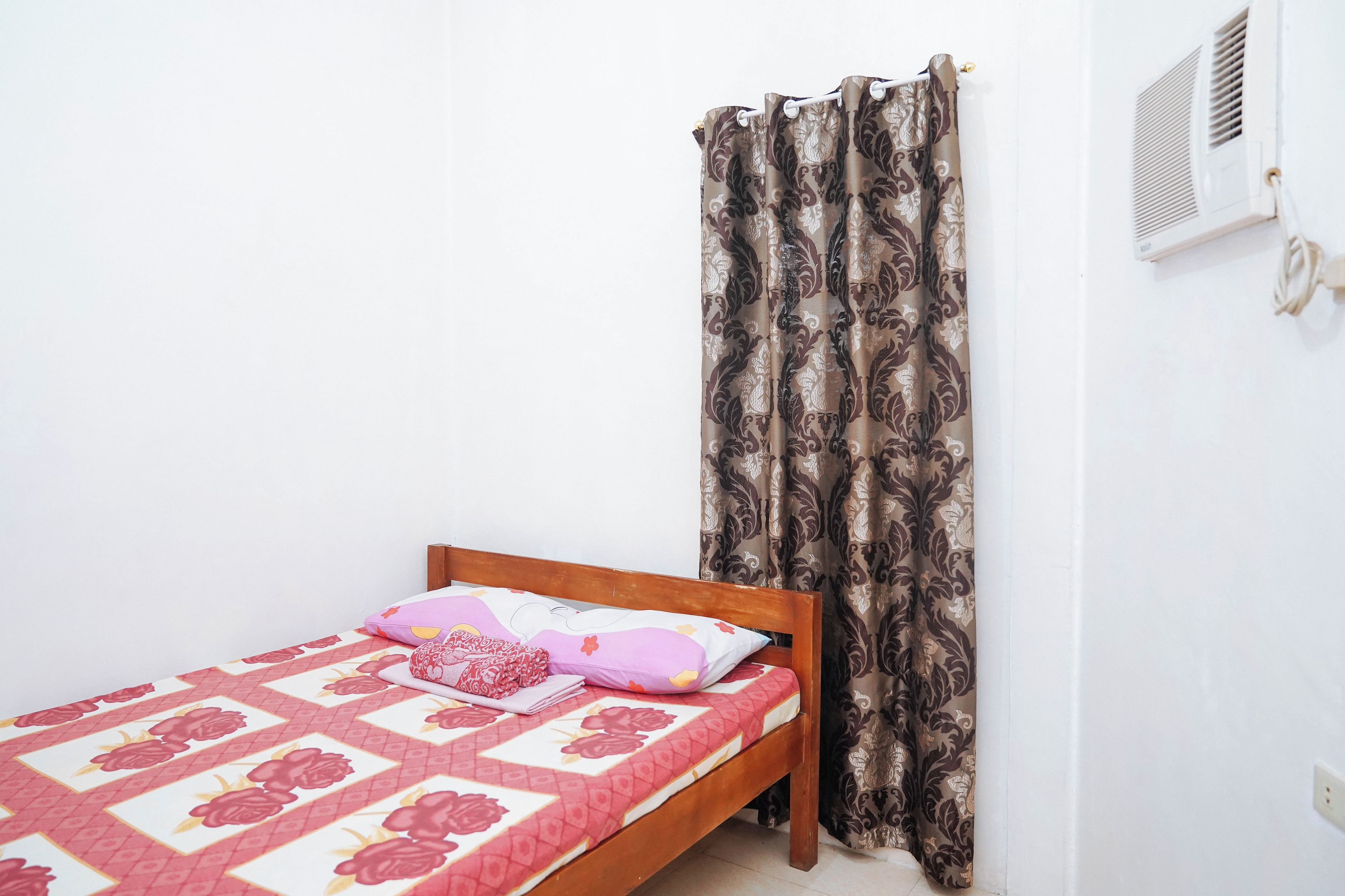 A single bed with a floral-patterned bedspread and a wooden headboard. A window with a white air conditioning unit is visible to the right.