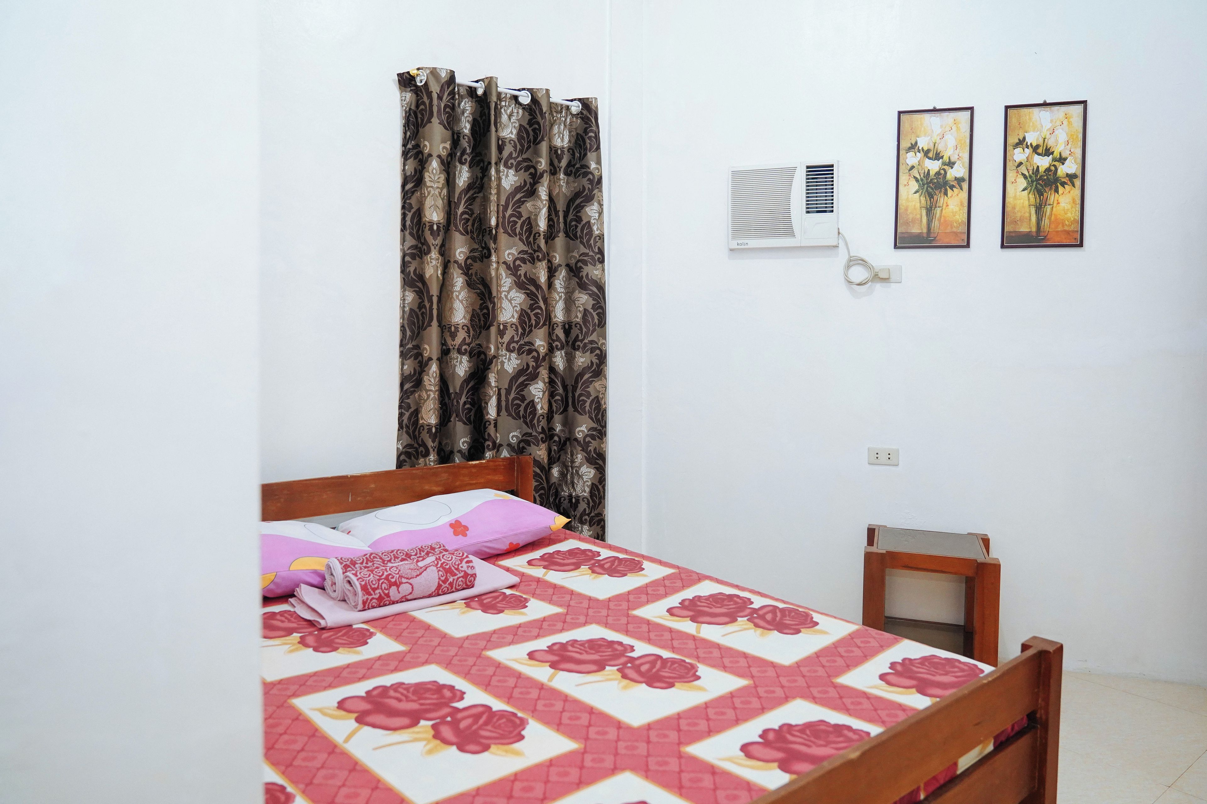 A bedroom with a bed, a small bedside table, a wall-mounted air conditioner, and two framed pictures on the wall.