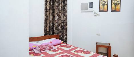 A bedroom with a bed, a small bedside table, a wall-mounted air conditioner, and two framed pictures on the wall.