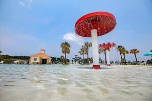 Una fuente de agua con parte superior roja y base blanca, rodeada de palmeras y un pequeño edificio con techo rojo.