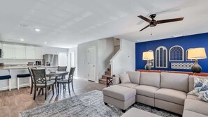 Un salon moderne avec un canapé d’angle beige, un coin repas comprenant une table et des chaises, et une cuisine avec des meubles blancs.