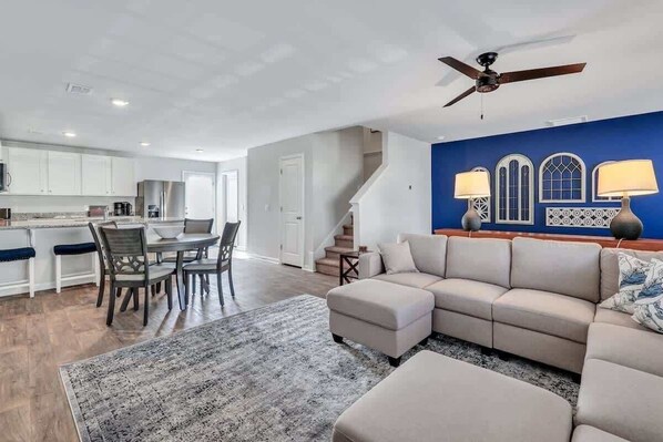 Una sala de estar moderna con un sofá seccional color beige, un comedor con mesa y sillas, y una cocina con armarios blancos.