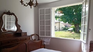 A bedroom with a wooden dresser, a chair, a bed with white bedding, a large mirror, and a window with a view of a garden and houses.
