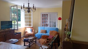 A living room with a television, a sofa, a chair, a table with a lamp, and a window with a view of greenery.