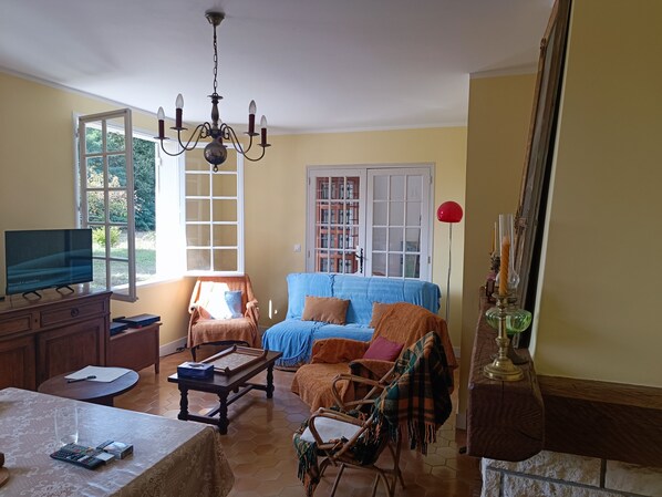 A living room with a television, a sofa, a chair, a table with a lamp, and a window with a view of greenery.