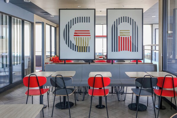 Una cafetería moderna con sillas rojas y negras, mesas blancas y arte abstracto en la pared.