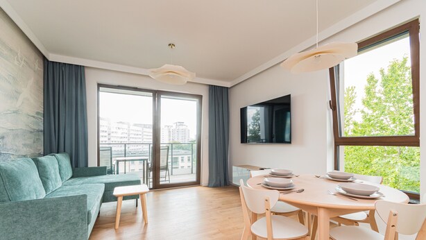 A modern living room with a green sofa, a wooden dining table with chairs, and a large window offering a city view. - Bulvar Premium by Grand Apartments (Gdansk)