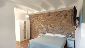 A bedroom with a stone wall, a bed with a light blue bedspread, two bedside tables with lamps, and a wooden chair. - Rincón de Piedra a 30' de Barcelona (Corró d'Avall)