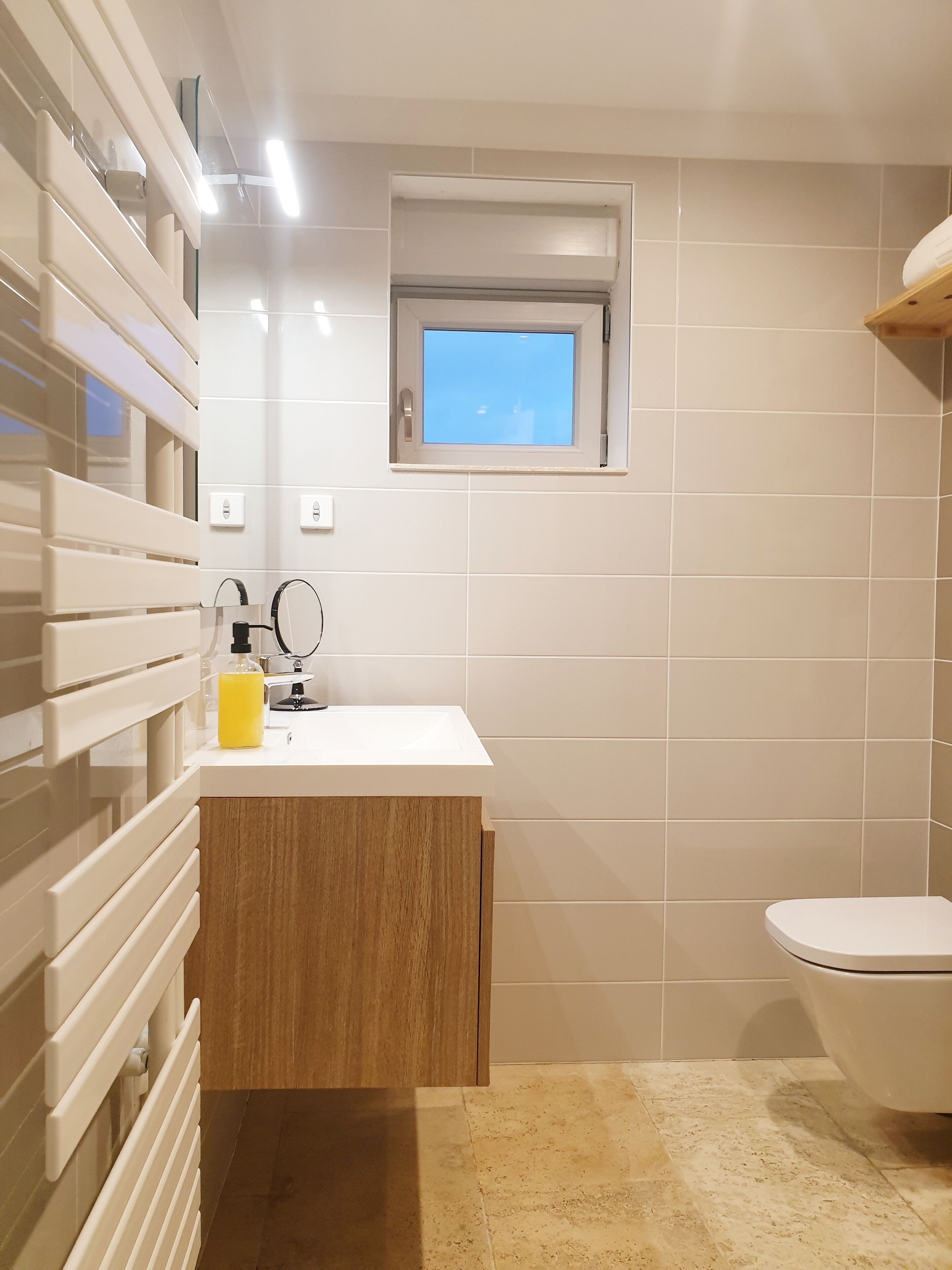 Une salle de bain moderne avec un lavabo blanc, un meuble en bois et une petite fenêtre située au-dessus du miroir.