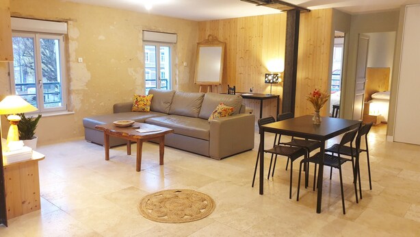 A living room with a sofa, coffee table, dining table, and chairs. There is a window, a lamp, and a plant. - Un Appart Dans le Perche, Votre Pied à Terre ! (Nogent-le-Rotrou)