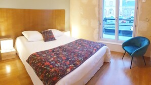 A hotel room with a bed, a chair, and a window with a view of buildings. - Un Appart Dans le Perche, Votre Pied à Terre ! (Nogent-le-Rotrou)