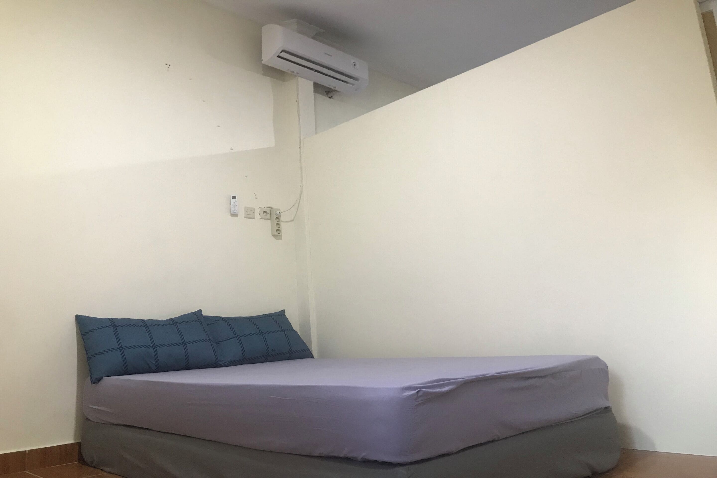 A small, single-bed room with a grey mattress, a blue pillow, and an air conditioning unit mounted on the wall.