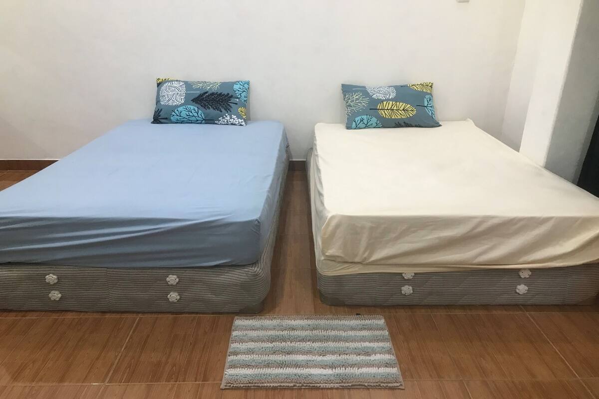 Zwei Einzelbetten mit blumigem Kissen, ein Streifen Teppich und eine schlichte Wand.