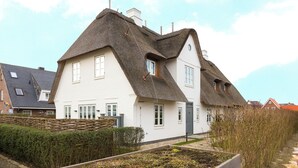 A white house with a steeply pitched thatched roof, surrounded by a neatly trimmed hedge and a wooden fence. - Part of the house in the basement, ground floor, first floor, approx. 200 sqm floor space, non-smoking, no pets (Wenningstedt-Braderup (Sylt))