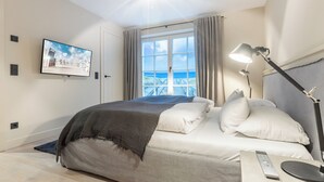 A bedroom with a bed, a TV mounted on the wall, a window with curtains, and a lamp on the bedside table. - Part of the house in the basement, ground floor, first floor, approx. 200 sqm floor space, non-smoking, no pets (Wenningstedt-Braderup (Sylt))