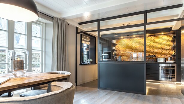 A modern kitchen with a brick wall backsplash, a dining area with a wooden table, and a large window with curtains. - Part of the house in the basement, ground floor, first floor, approx. 200 sqm floor space, non-smoking, no pets (Wenningstedt-Braderup (Sylt))