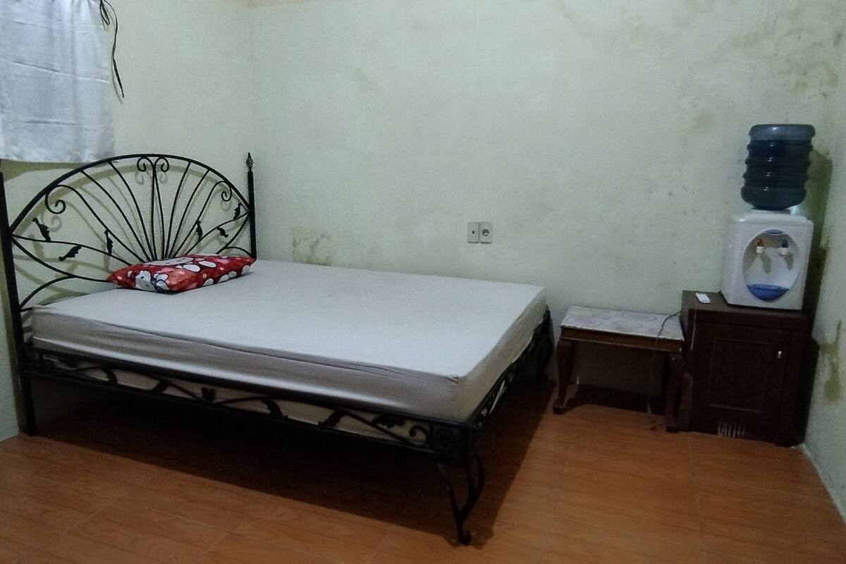 Une petite chambre avec un lit simple, un cadre de lit en métal, une table de chevet, un distributeur d’eau et une prise électrique au mur.