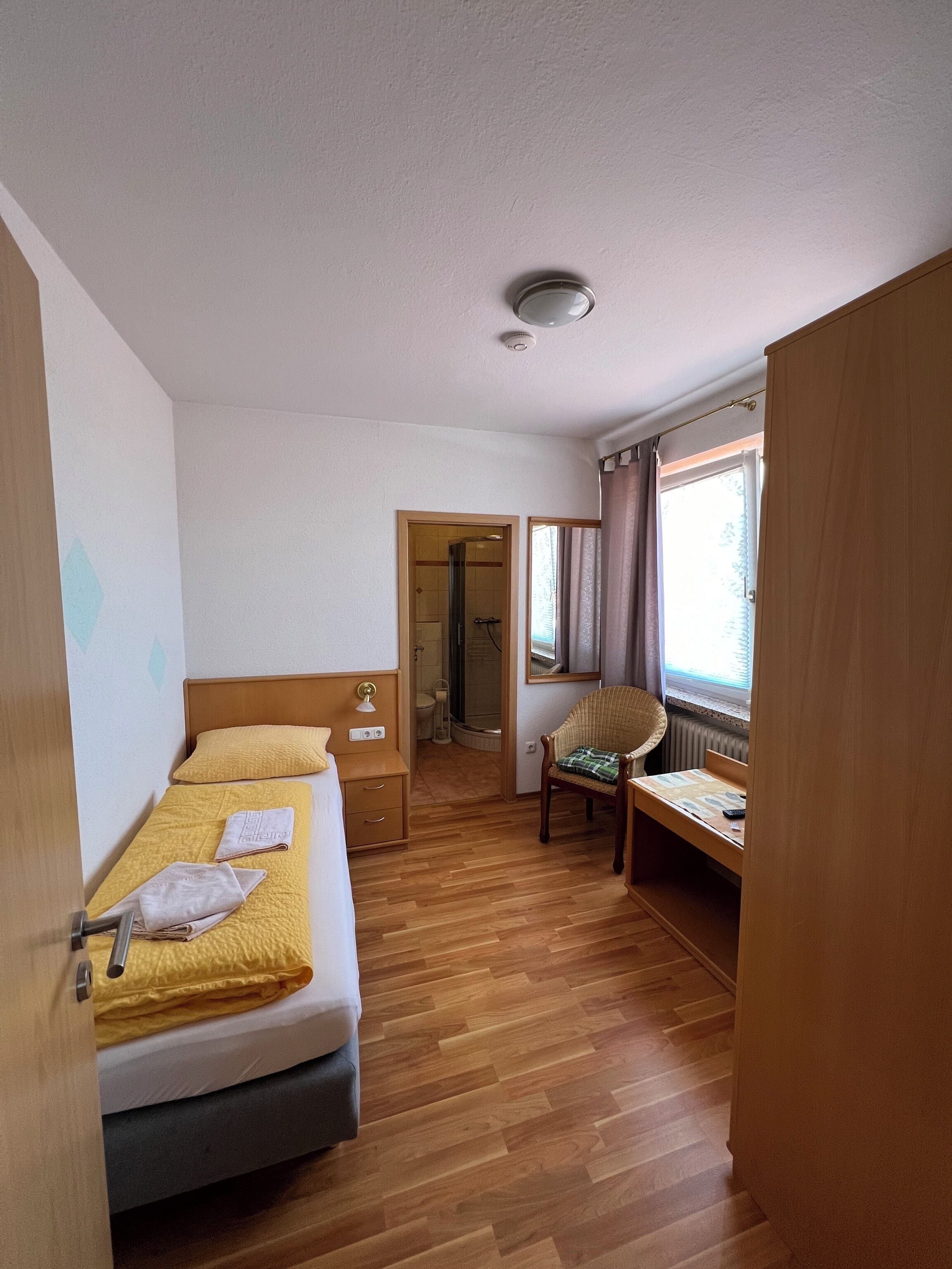 Ein kleines Hotelzimmer mit Bett, Schreibtisch, Stuhl und einem durchgegangenen Tür sichtbaren Badezimmer.