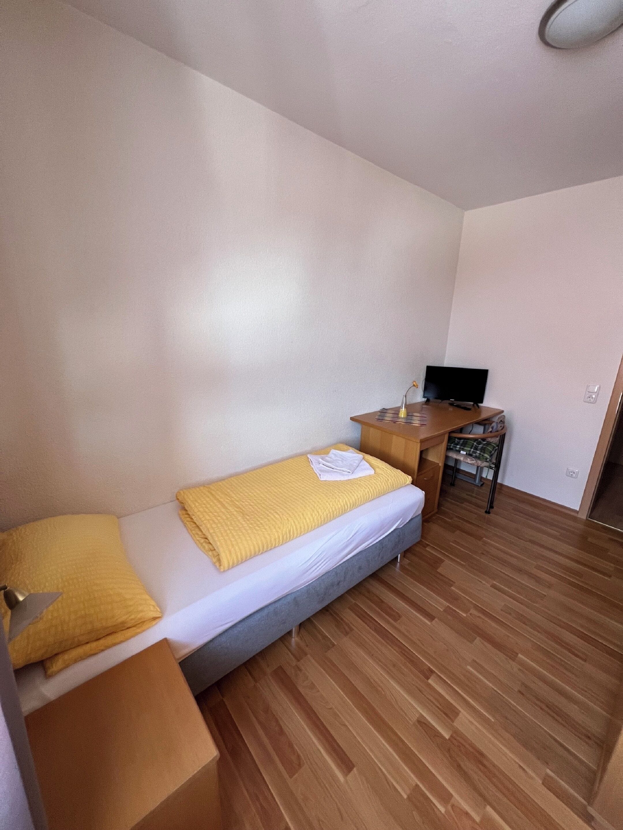 Una habitación pequeña y ordenada con cama, escritorio y televisión.