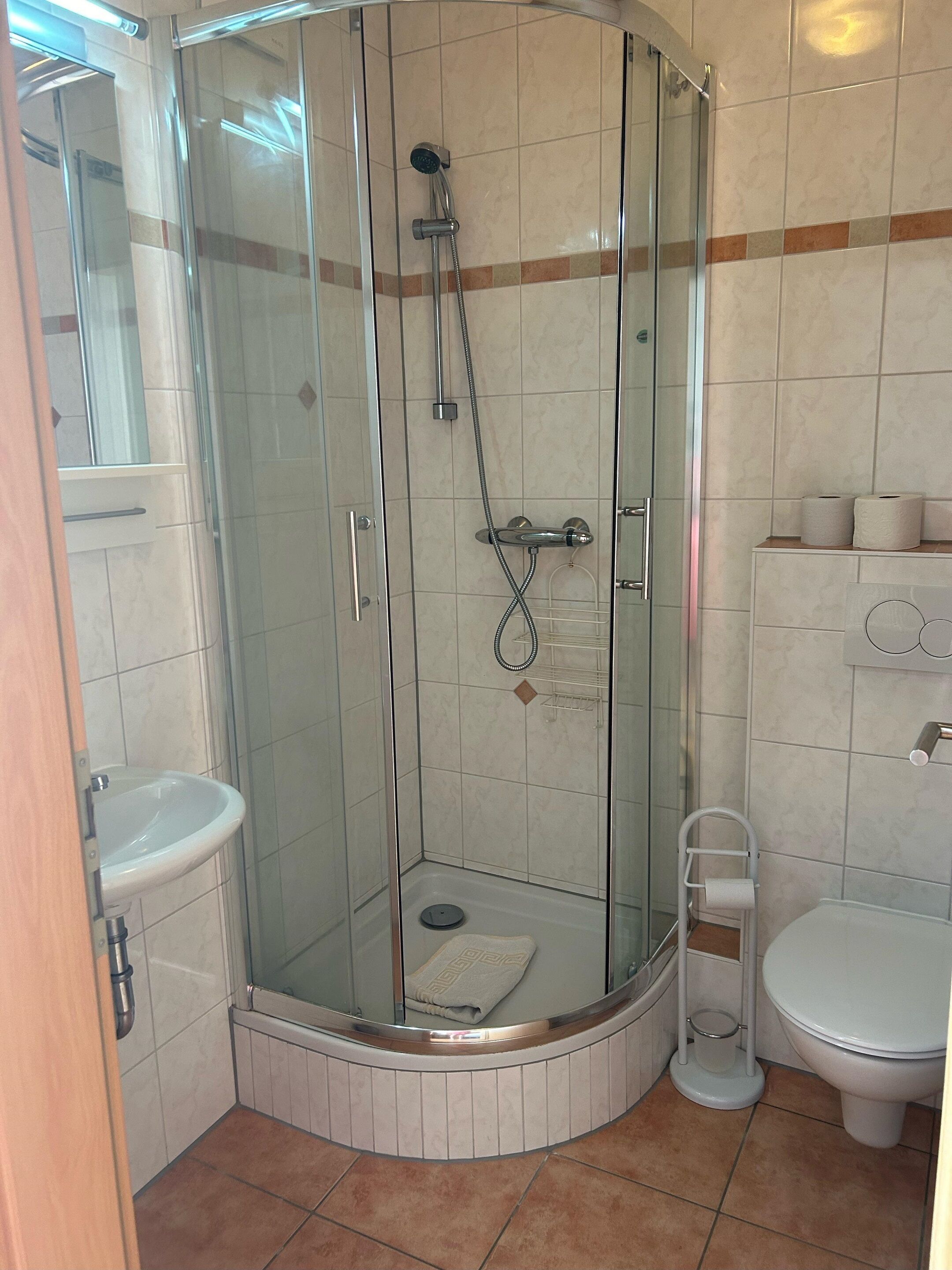 Baño en esquina con ducha de mampara de vidrio, lavamanos e inodoro.