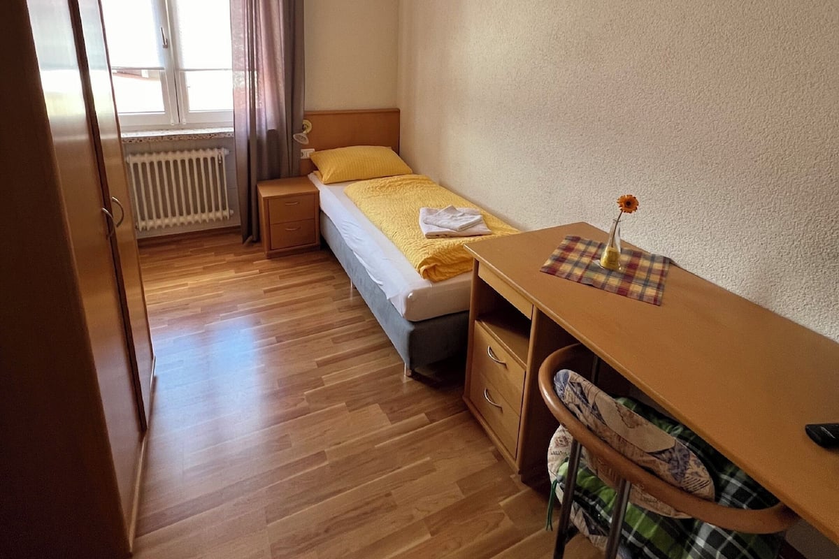 Een kleine, verzorgde slaapkamer met een enkel bed, een houten bureau, een stoel en een radiator.