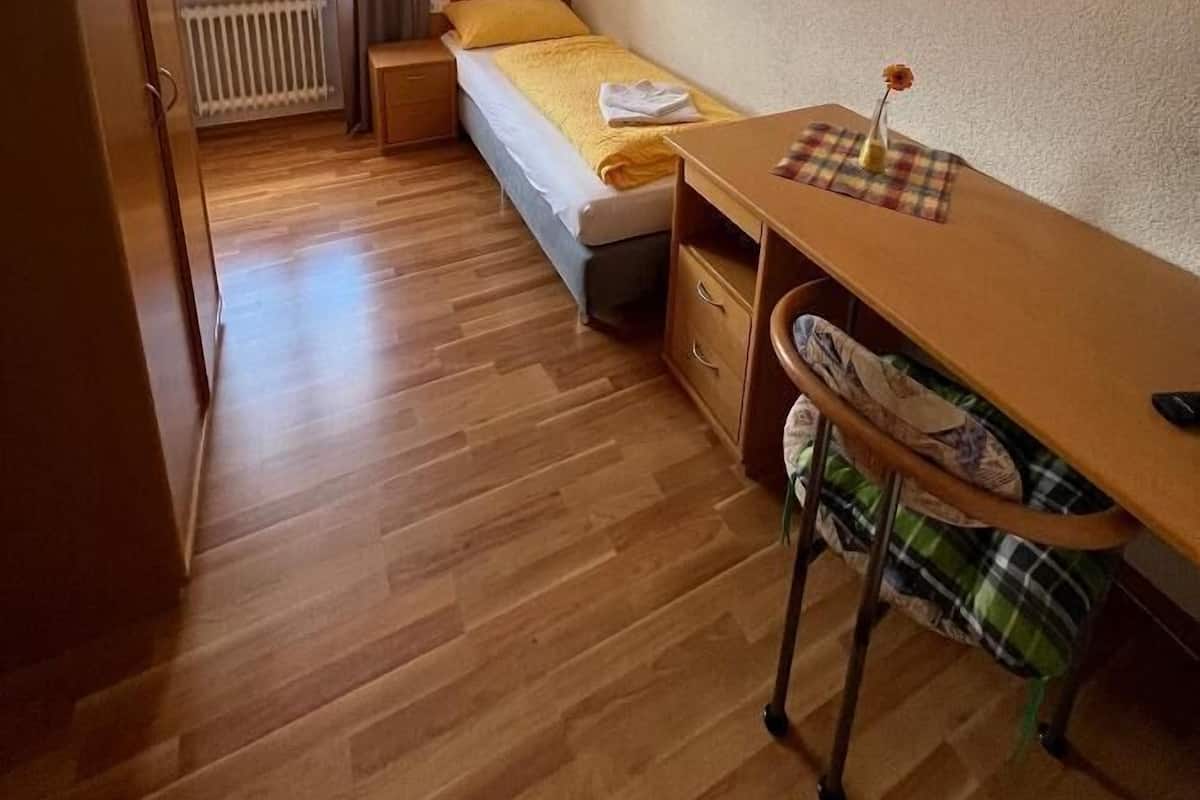Een kleine eenpersoonskamer met een houten bureau, een stoel, een radiator en een raam met gordijnen.
