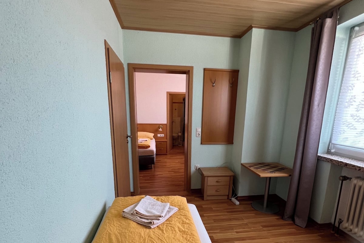 Een kamer met een bed, een klein tafeltje, een nachtkastje, een raam met gordijnen en een deur naar een andere kamer.