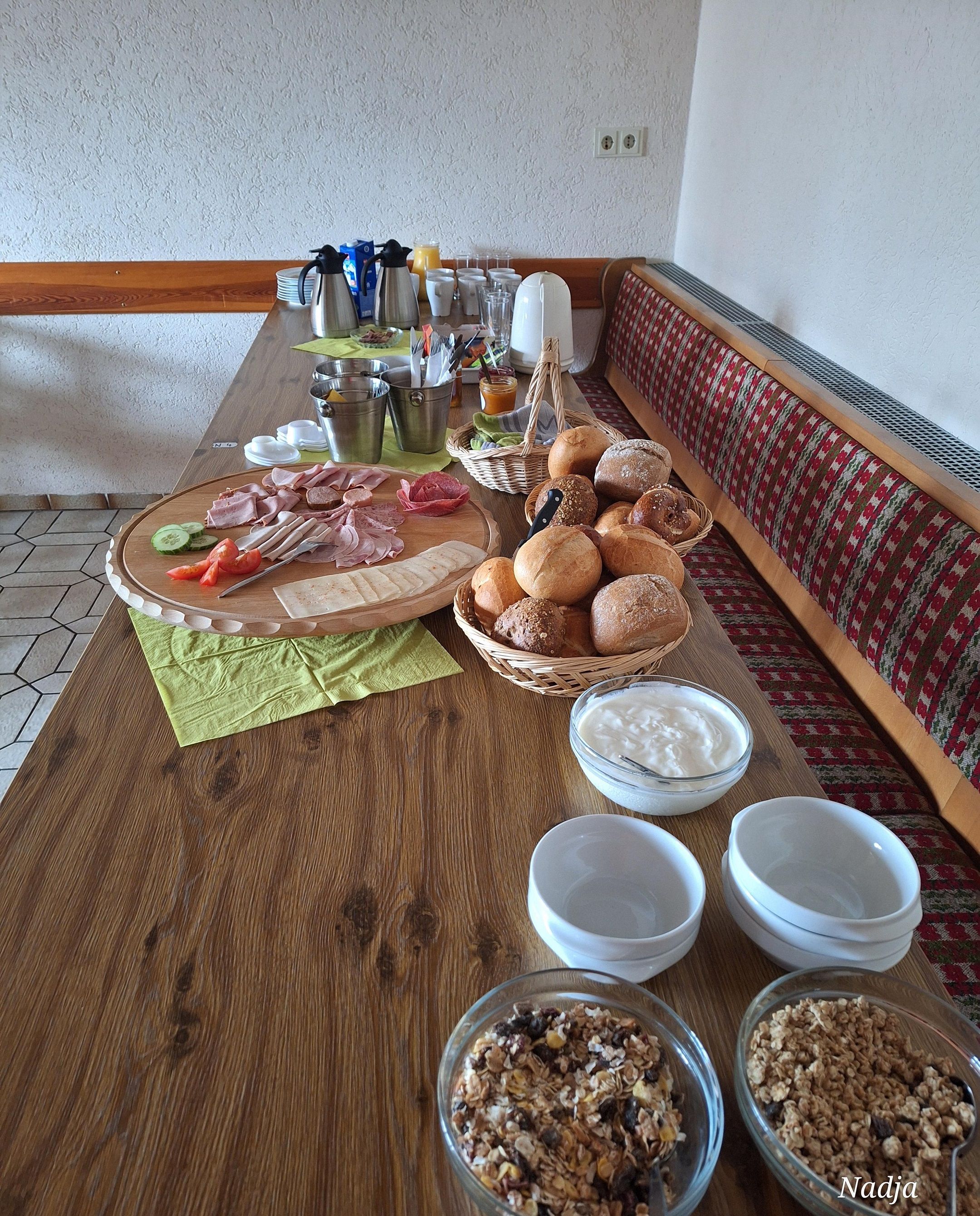 Una mesa de madera con desayuno servido, incluyendo panes variados, una tabla de fiambres, tazones de yogur y granola.