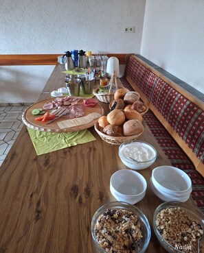 Disposição para o pequeno-almoço com uma mesa de madeira, pães variados, uma tábua de enchidos e taças de iogurte e granola.