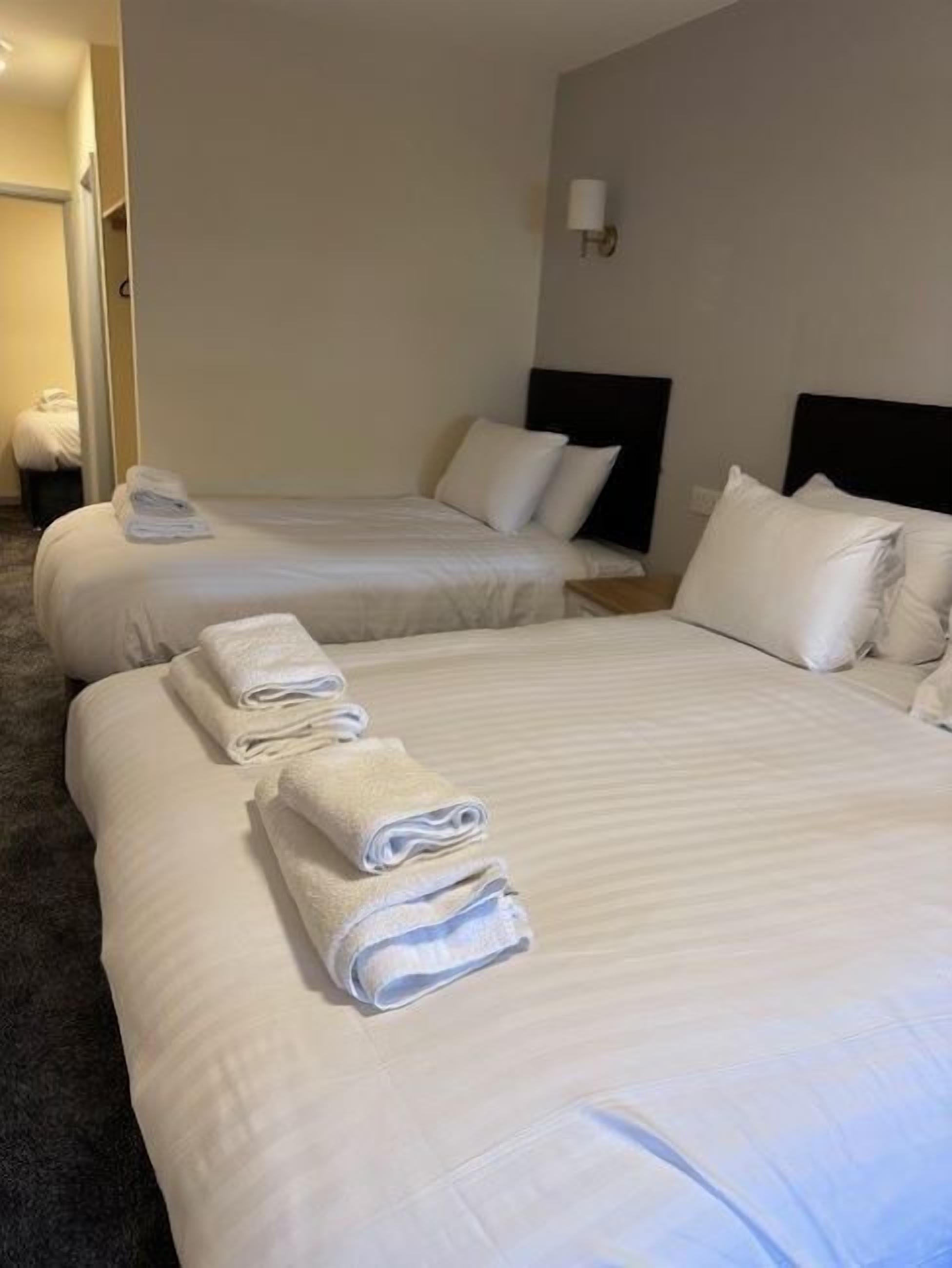 A hotel room with two beds, each with white linens and pillows, and a small bedside table with a lamp.