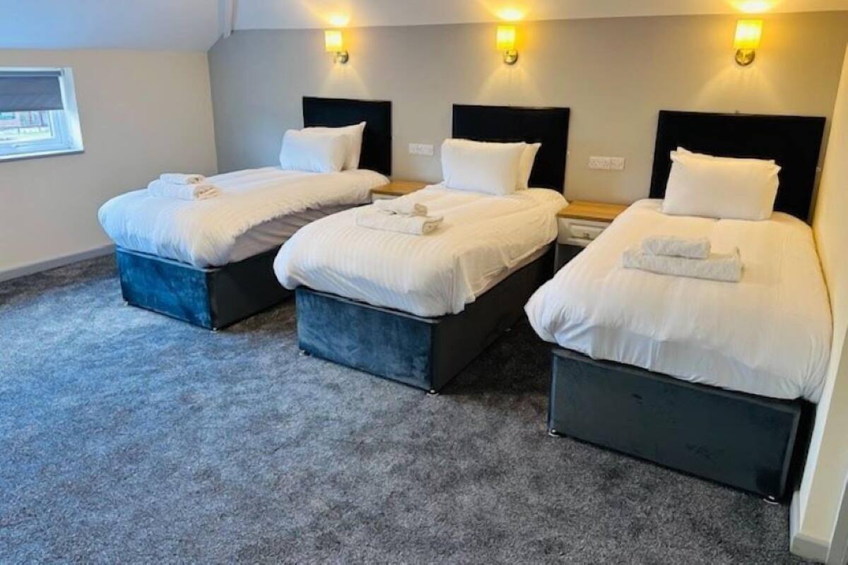 A hotel room with three single beds, each with a dark headboard and white bedding. There are wall-mounted light fixtures above each bed.