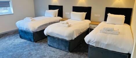 A hotel room with three single beds, each with a dark headboard and white bedding. There are wall-mounted light fixtures above each bed.